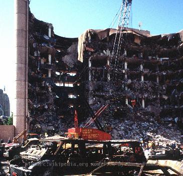 The bombed remains of automobiles with the bombed Federal Building in the background. The military is providing around the clock support since a car bomb exploded inside the building on Wednesday, April 19, 1995. (Released to Public)