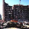 The bombed remains of automobiles with the bombed Federal Building in the background. The military is providing around the clock support since a car bomb exploded inside the building on Wednesday, April 19, 1995. (Released to Public)